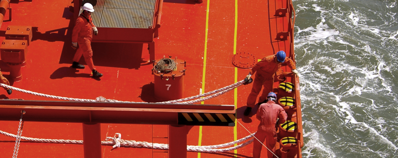 Drei Matrosen in orangener Schutzkleidung und Helmen arbeiten an Deck eines Schiffes mit dicken Tauen am Meer.